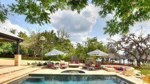 A swimming pool with lounge chairs and umbrellas, surrounded by trees and a wooden fence. - LOCATION!!  Lake Travis Waterfront~Pool, Spa, Outdoor Kitchen, Location (Austin)
