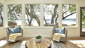 A room with large windows overlooking a body of water, wicker furniture, and a round wooden table with a plant. - LOCATION!!  Lake Travis Waterfront~Pool, Spa, Outdoor Kitchen, Location (Austin)