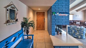 Un pasillo con una puerta de madera, un muro de ladrillo azul, un cartel que dice "Welcome to Paradise", un sofá azul y una pequeña mesa con objetos decorativos.