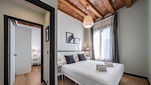 A modern bedroom with a large bed, wooden flooring, a ceiling with exposed beams, and a large window with curtains.