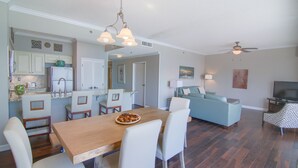 Una sala de estar amplia con un sofá azul, un comedor con una mesa de madera y sillas blancas, y una cocina con armarios blancos y un refrigerador.