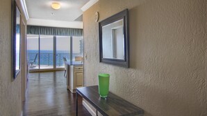 Un pasillo con suelo de madera, un jarrón de vidrio verde sobre una mesa, un espejo en un marco en la pared y vista al océano a través de una puerta de balcón.