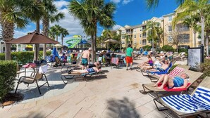 A poolside area with lounge chairs, palm trees, and a water slide. - Disney Magic Awaits: 5-BDRM, 3-Suites, Pool, Upgraded Perks,  and Tile Flooring! (Kissimmee)