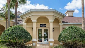 A building with a 'Welcome Center' sign, a covered entrance, and symmetrical landscaping. - Disney Magic Awaits: 5-BDRM, 3-Suites, Pool, Upgraded Perks,  and Tile Flooring! (Kissimmee)