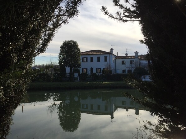 A two-story building with a tiled roof, surrounded by trees and a calm body of water reflecting the structure.