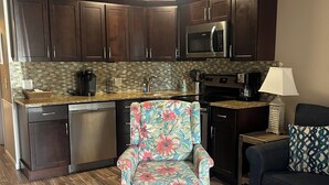 A compact kitchen with dark wooden cabinets, a microwave, and a coffee maker. A floral armchair is placed near the kitchen, and a sofa with a patterned pillow is in the living area.