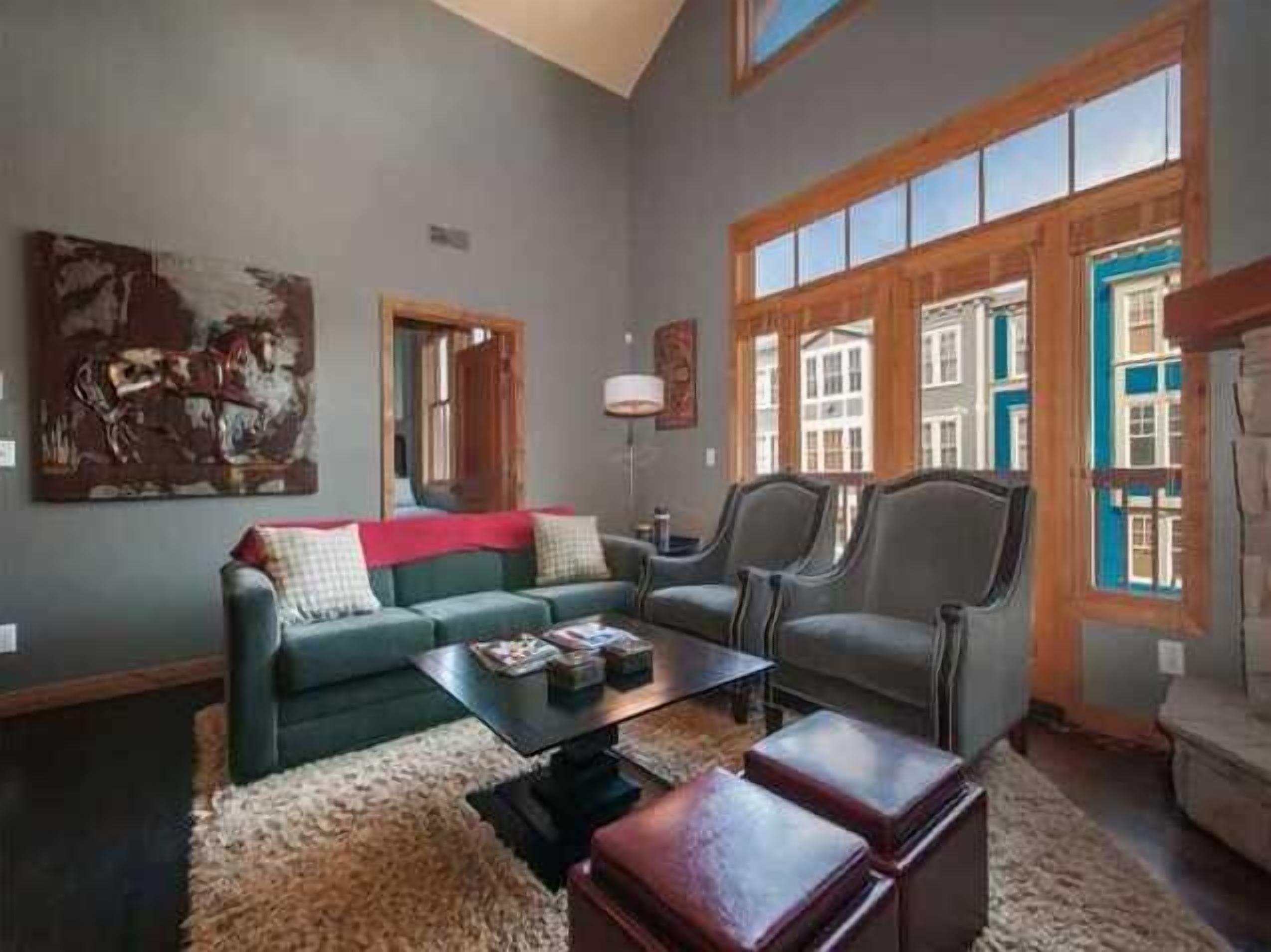Un salon comprenant un canapé, des fauteuils, une table basse et un tapis. Un tableau est accroché au mur, et une fenêtre offre une vue sur les bâtiments.