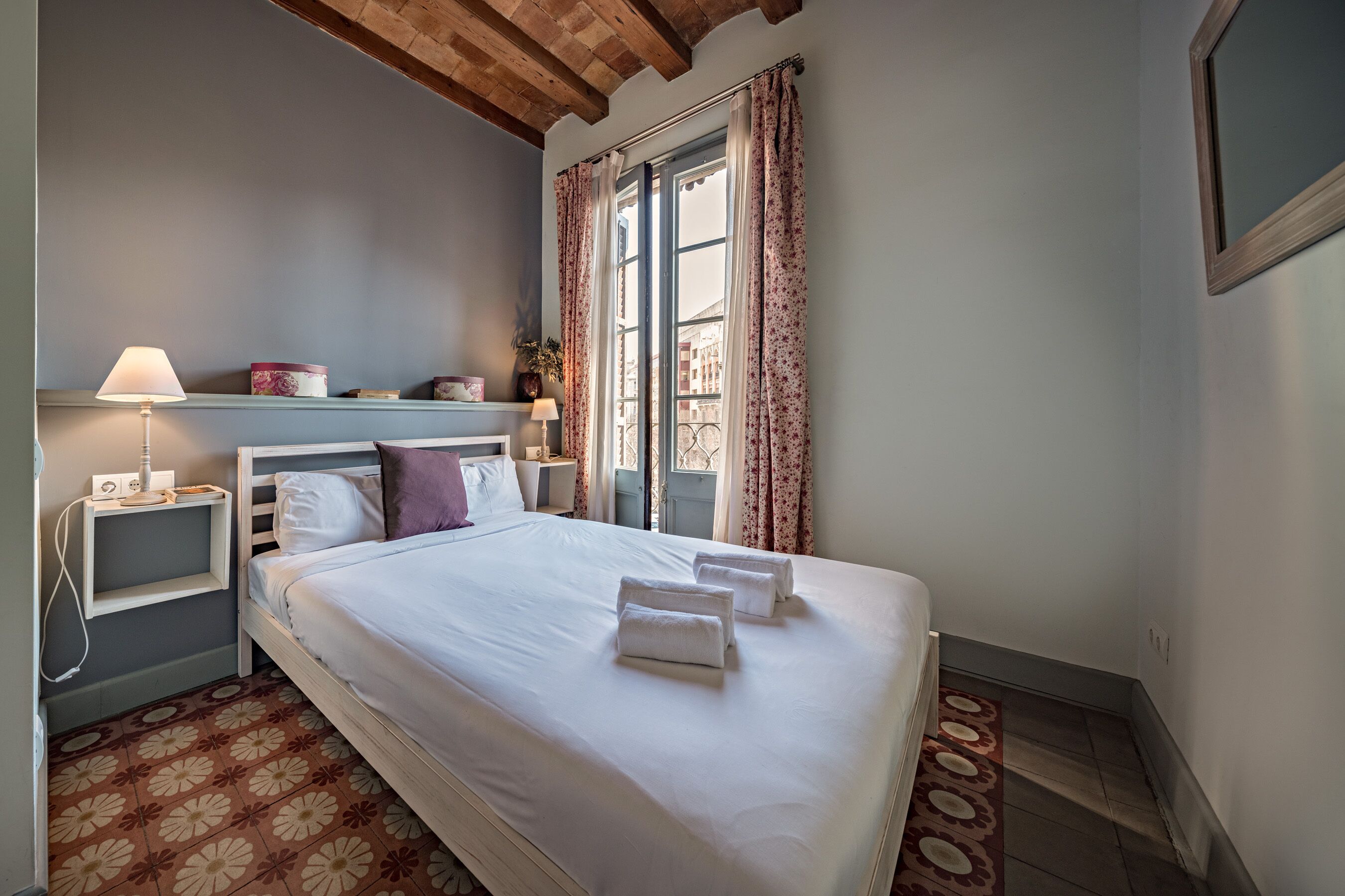 A neatly made bed with white linens and towels, a bedside lamp, a nightstand, and a window with floral curtains.