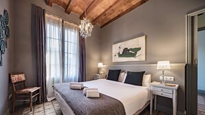 Habitación de hotel ordenada con una cama grande, techo de madera, candelabro, un cuadro, una silla y una mesita de noche con lámpara.