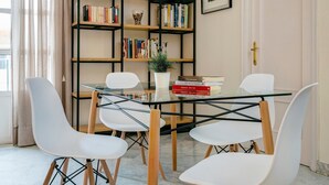 A dining area with a glass table, white chairs, a bookshelf with books and decorative items, and a door with a brass handle. - Genteel Home Tomas de Ibarra (Sevilla)