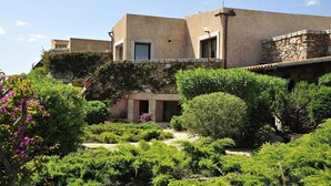 Um edifício com fachada de pedra, telhado plano e um jardim com arbustos e flores variadas.
