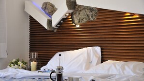 Quarto de hotel moderno com cabeceira de madeira, cama com roupa branca, máquina de café, um copo de água e uma vela sobre a mesa de cabeceira.