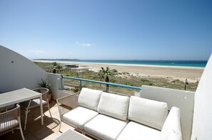 Un balcon avec un canapé et des fauteuils blancs, une petite table et une plante en pot. Le balcon donne sur une plage, et le ciel est dégagé.