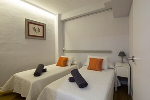 A hotel room with two beds, each with a white bedspread and two pillows, one orange and one white. There is a framed picture on the wall, a bedside table with a lamp, and a wooden headboard.