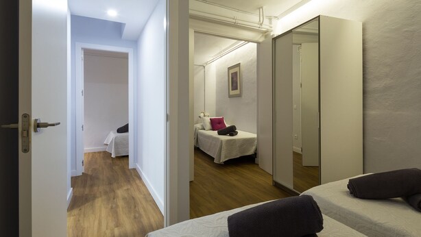 Una camera d'albergo con un letto, un comodino, una sedia e una porta che dà su un'altra stanza.