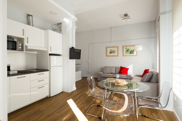 A modern kitchen with white cabinets, a microwave, and a refrigerator. There is a dining area with a glass table and chairs. A sofa with red cushions is in the living area.