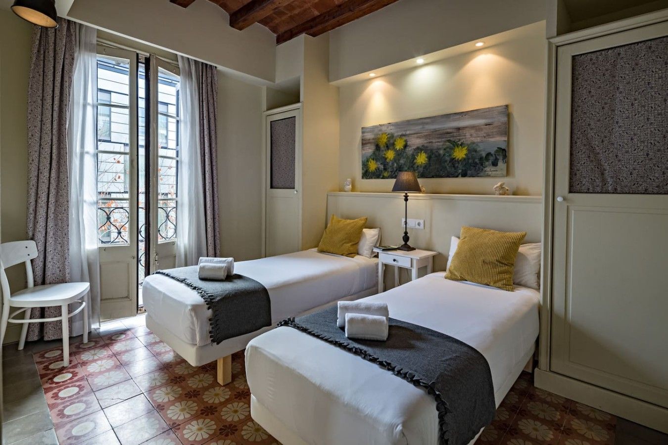Une chambre d’hôtel avec deux lits, une chaise, une fenêtre avec des rideaux, un tableau au mur et une lampe.