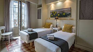 Une chambre d’hôtel avec deux lits, une chaise, une fenêtre avec des rideaux, un tableau au mur et une lampe.
