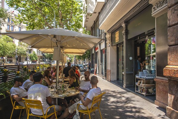 Ein Straßencafé mit gelben Stühlen und einem großen Sonnenschirm, umgeben von Gebäuden und Bäumen.