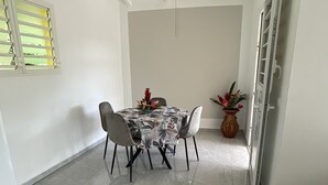 A dining area with a round table, four chairs, a floral tablecloth, a hanging light fixture, and a potted plant.