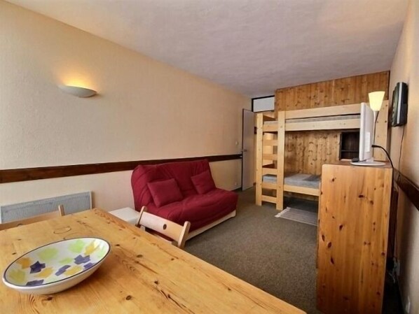 A compact room with a bunk bed, a red sofa, a wooden table, and a wall-mounted lamp.
