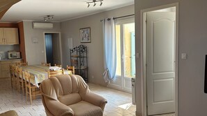 Un salón con un sillón color beige, una mesa de centro de madera y un comedor con mesa y sillas. Se observa una ventana con cortinas, una puerta y un equipo de aire acondicionado instalado en la pared.
