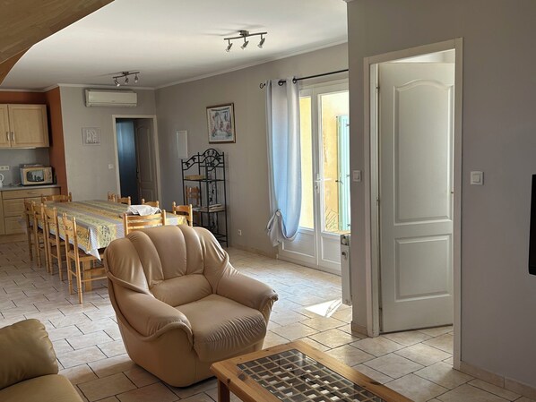 Een woonkamer met een beige fauteuil, een houten salontafel en een eethoek met tafel en stoelen. Er is een raam met gordijnen, een deur en een wandgemonteerde airconditioning.