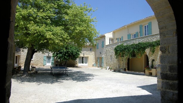 Un patio con un banco, un árbol y edificios de piedra con contraventanas azules.