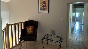 Una habitación con una silla, una mesita con lámpara, un cuadro enmarcado en la pared y un techo con vigas de madera.