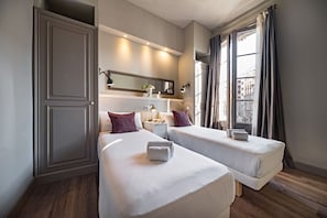 A hotel room with two beds, a wardrobe, a bedside table with lamps, and a window with curtains.