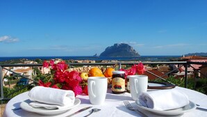 Une table dressée avec des assiettes, des couverts, un pot de confiture et une tasse, donnant sur une vue côtière avec une montagne imposante à l’horizon.