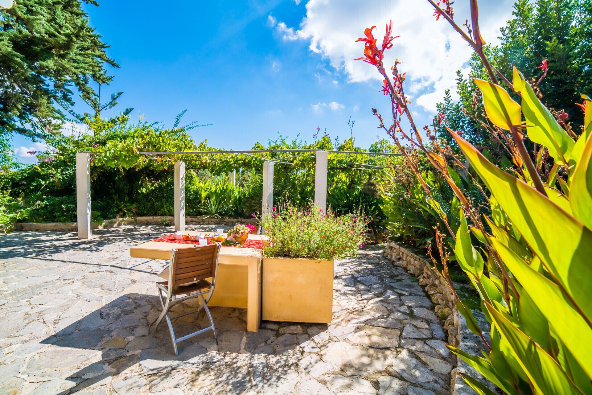 An outdoor patio with stone flooring, a wooden table and chair, and a pergola with climbing plants.