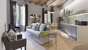 A modern living room with a sofa, armchair, and a small round table. There is a kitchen area with a refrigerator, microwave, and a coffee machine. The room has wooden beams on the ceiling and large windows with curtains.