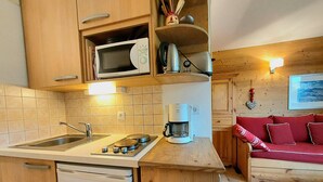 Een compacte keuken met een spoelbak, fornuis, magnetron en koffiezetapparaat. Daarboven bevindt zich een houten kastje en er is een rode bank in de woonkamer.