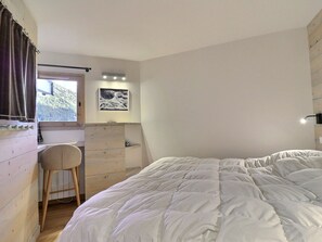 A bedroom with a bed, a desk, a chair, a window with curtains, a wall-mounted light, and a picture.