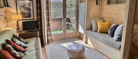 A cozy room with a sofa, a television, a window with curtains, and a view of a mountain.