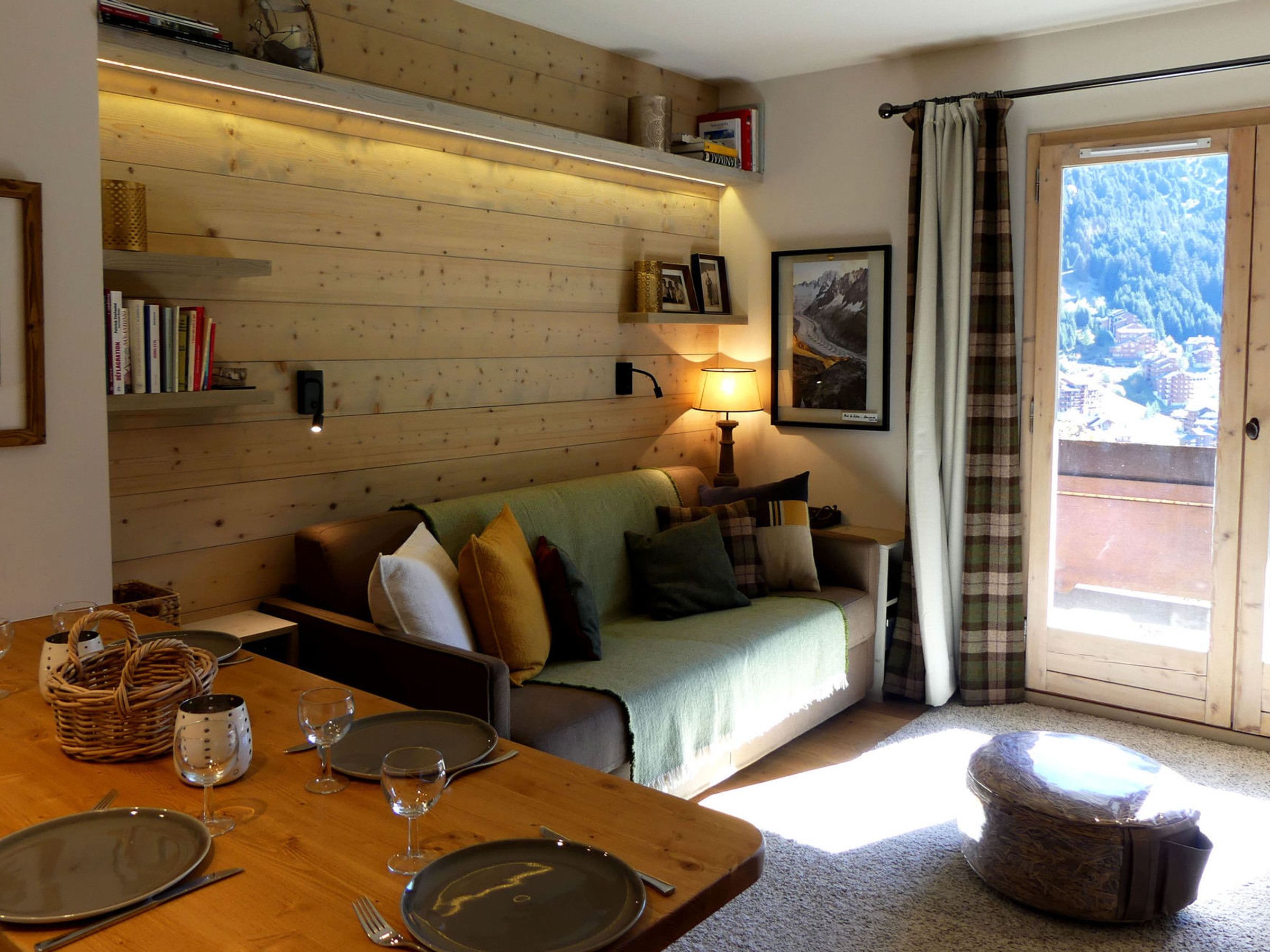 A cozy living room with a wooden wall, a sofa, a dining table set with plates and glasses, and a window with a view of a mountainous landscape.