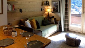 A cozy living room with a wooden wall, a sofa, a dining table set with plates and glasses, and a window with a view of a mountainous landscape. - 3-Room Apartment on the Slopes, Perfect for Families (Méribel)