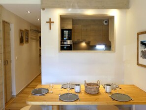 A dining area with a wooden table set for two, a kitchen visible through an archway, and a cross on the wall.