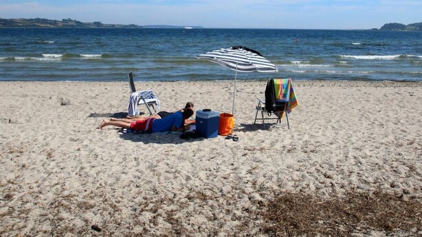 A beach with a person lying on a towel under a striped umbrella, a chair, and a cooler. - Beautiful holidayhouse with seaview and wellness area (Broager)