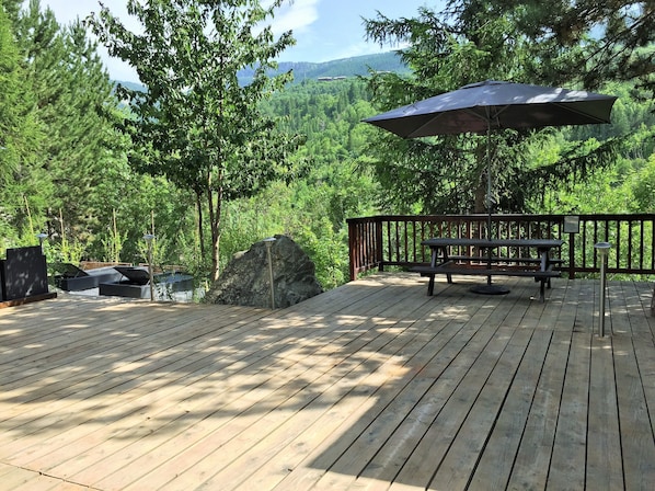 Una terraza de madera con mesa de picnic y sombrilla, rodeada de árboles y vistas a montañas.