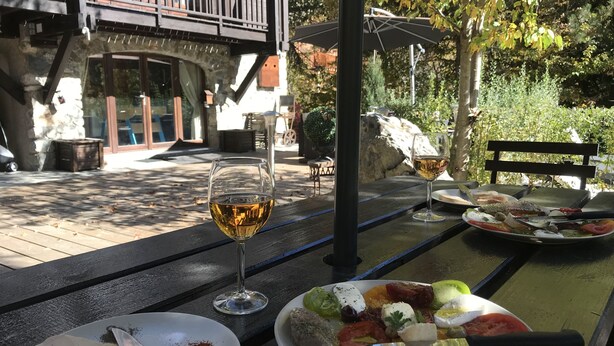 Ein Essbereich im Freien mit Tisch, gedeckten Plätzen, einem Weinglas und Blick auf ein Steinhaus mit Balkon.