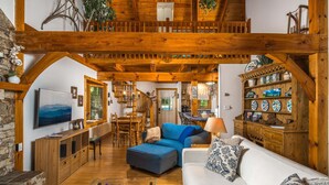 Un intérieur rustique avec un plafond en bois, un coin repas, un salon avec un canapé et un pouf, et une cuisine visible à travers une porte ouverte.