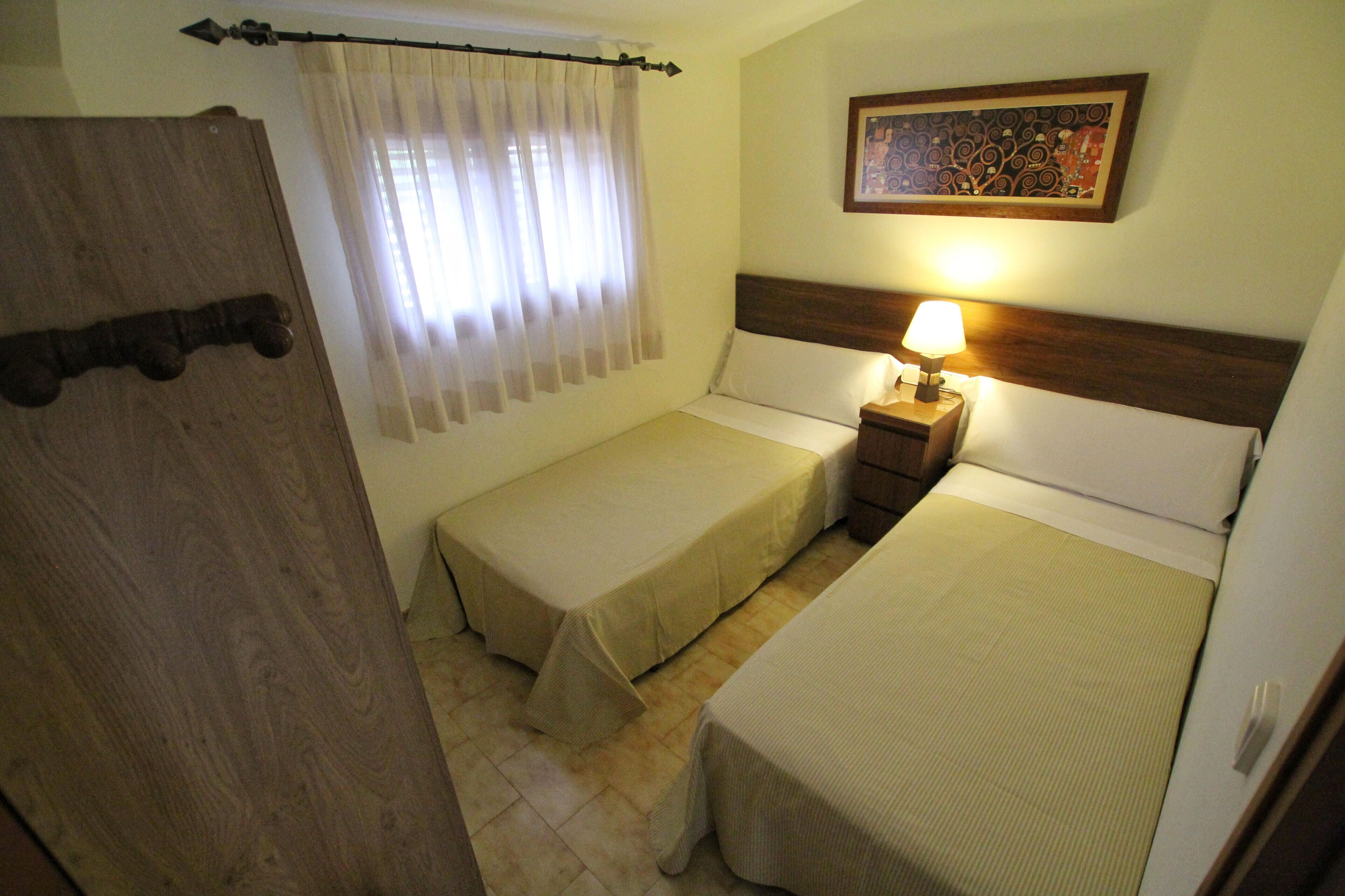 Una habitación de hotel pequeña con dos camas individuales, un cabecero de madera, una mesita de noche con una lámpara, una cortina y un cuadro en la pared.