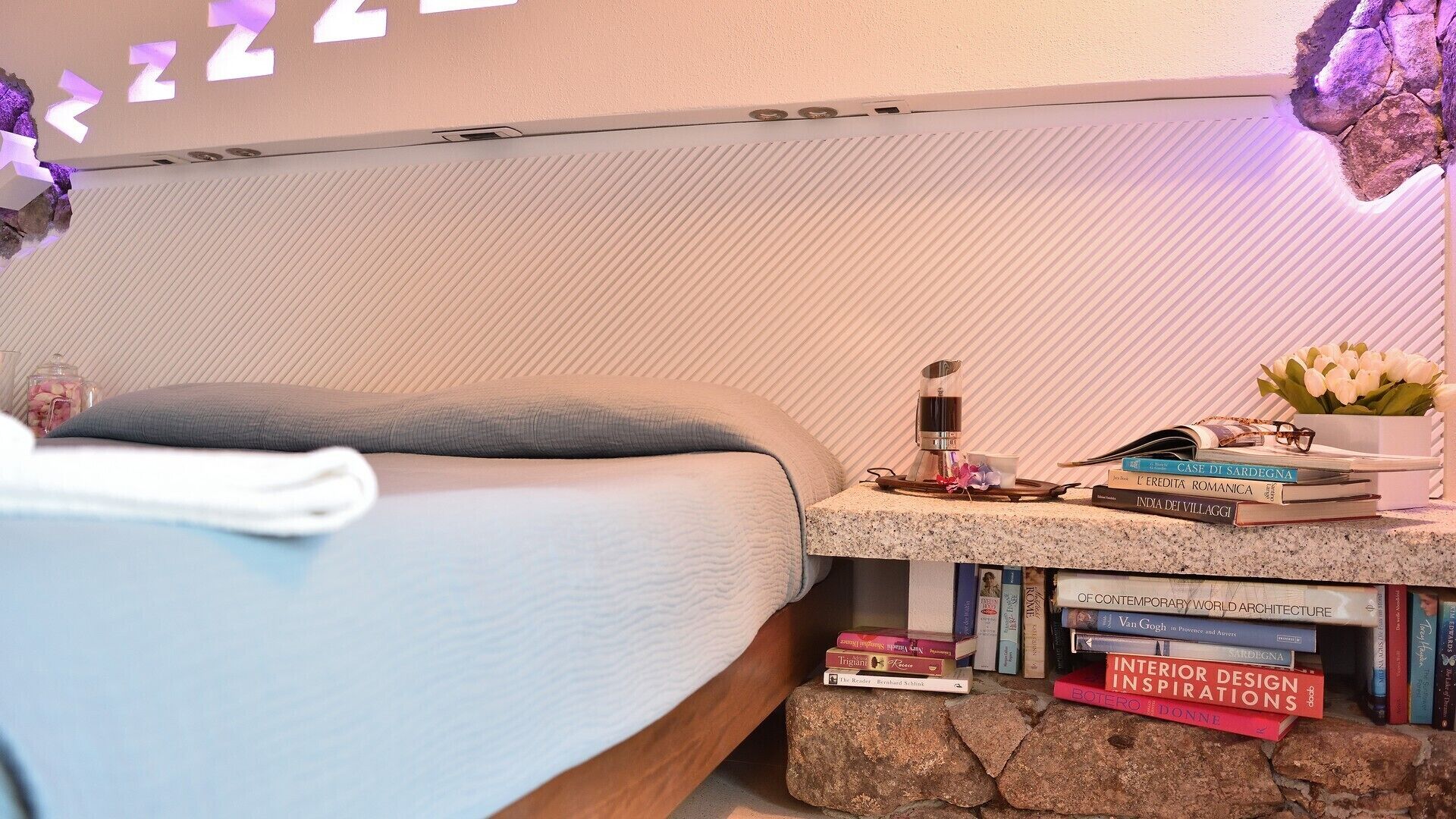 A neatly made bed with a grey blanket, a bedside table with books and a small plant, and a wall with a textured pattern.
