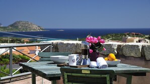 A table with a coffee pot, cups, a bowl of fruit, and a vase of flowers on a balcony overlooking the sea. - Holiday apartments L'Ea di Lavru 
SEA VIEW 2-rooms apartment-TOP INTERIOR DESIGN (Porto San Paolo)
