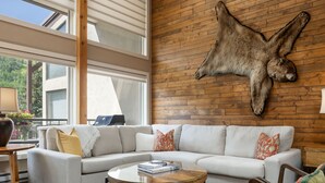 Ein modernes Wohnzimmer mit einer Eckcouch, einem runden Couchtisch und einem Ledersessel. Ein großer Fellteppich ist an der Holzwand angebracht, und auf der Couch liegt ein Laptop.