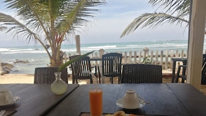A table set with a plate of fruit, a glass of orange juice, and a cup, overlooking a beach with palm trees and a wooden fence. - Happy Beach (Unawatuna)