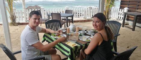 A couple dining at a table with a view of the ocean, surrounded by chairs and a white fence.