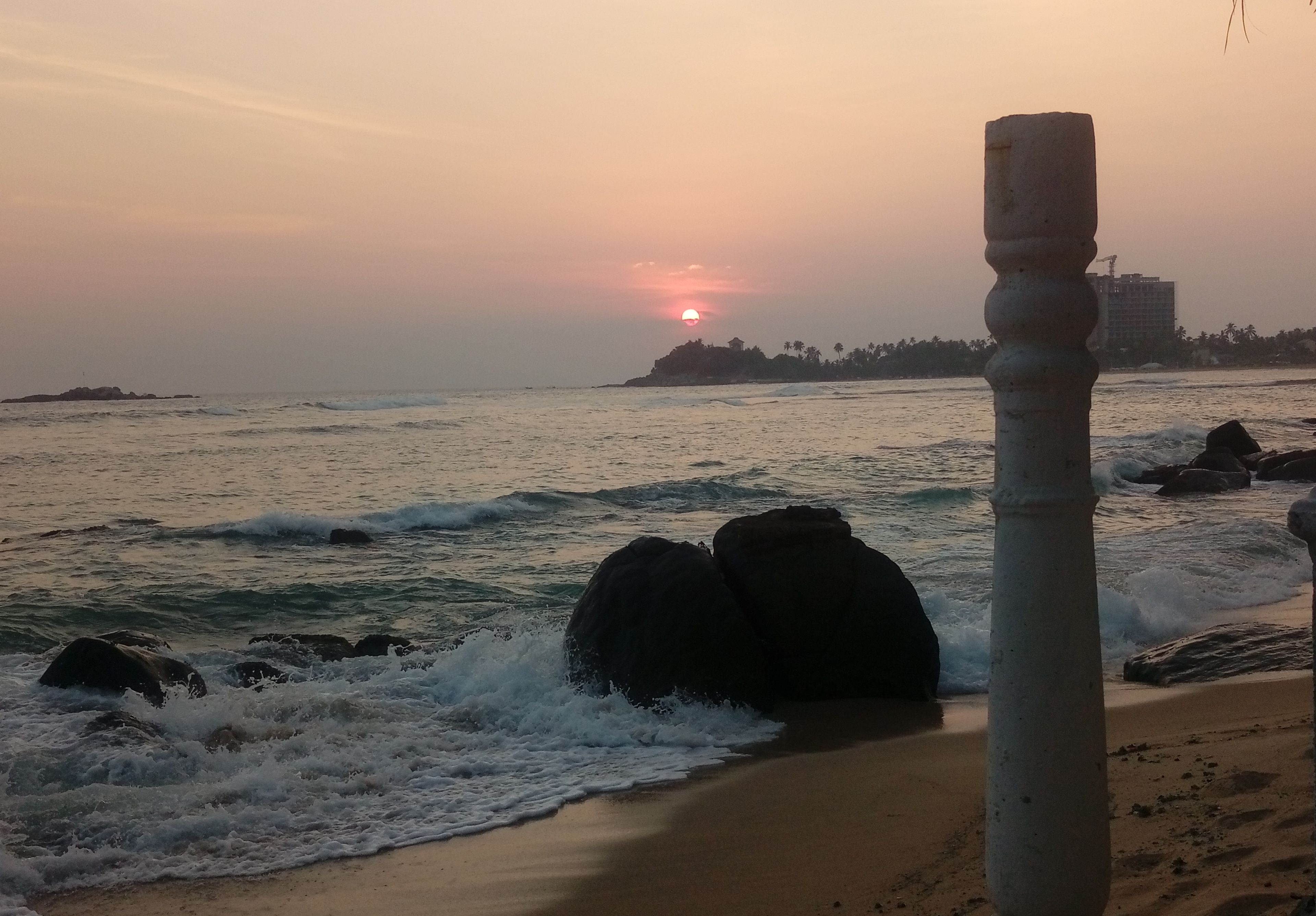A beach scene with waves, a sunset, and a stone pillar.